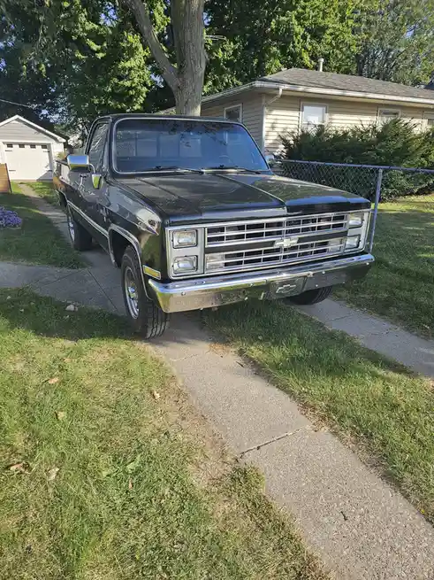 A black 1980s Chevrolet pickup truck is parked on a concrete driveway in a suburban yard, partially on grass. The truck has…