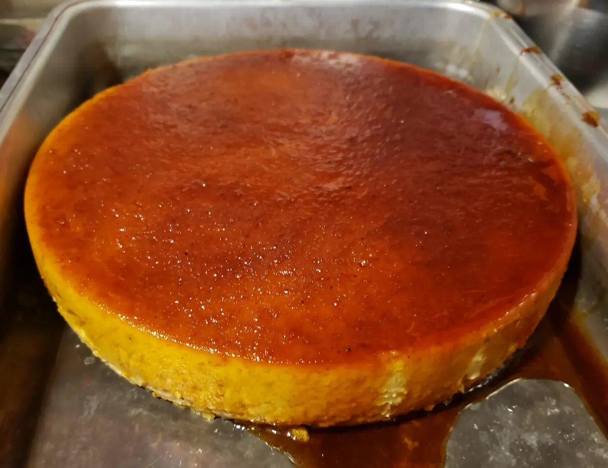 A close-up photo shows a round, golden-brown baked dessert in a metal baking pan, with a glossy caramelized top and some…