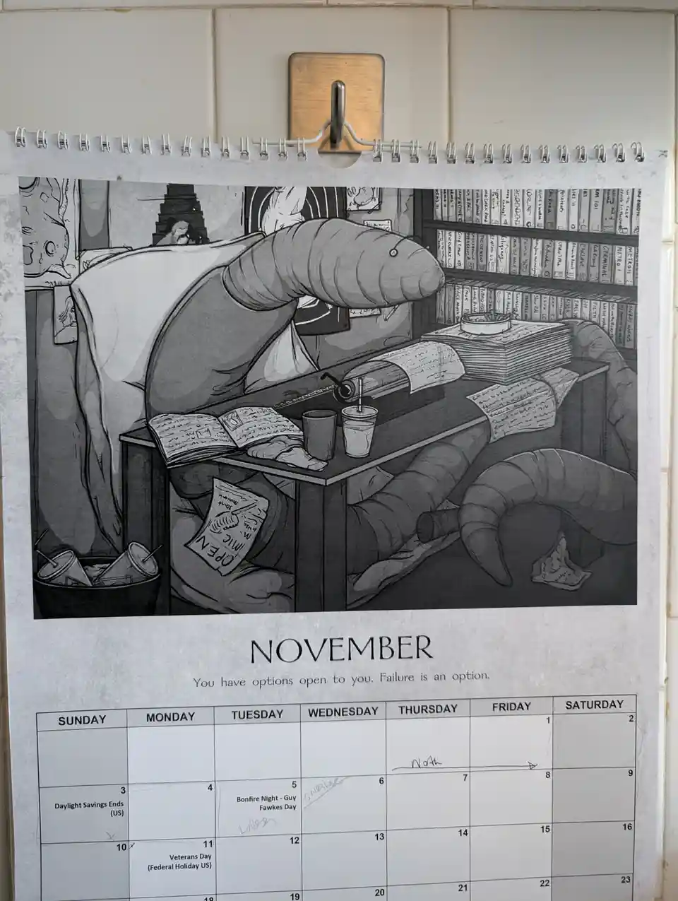 A black-and-white calendar page for November shows a cartoon worm-like creature slumped over a cluttered desk, surrounded by…