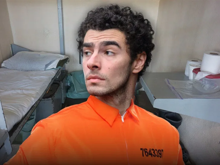 A man with curly dark hair and a skeptical expression wears an orange prison jumpsuit with the number 7643397 on the chest…