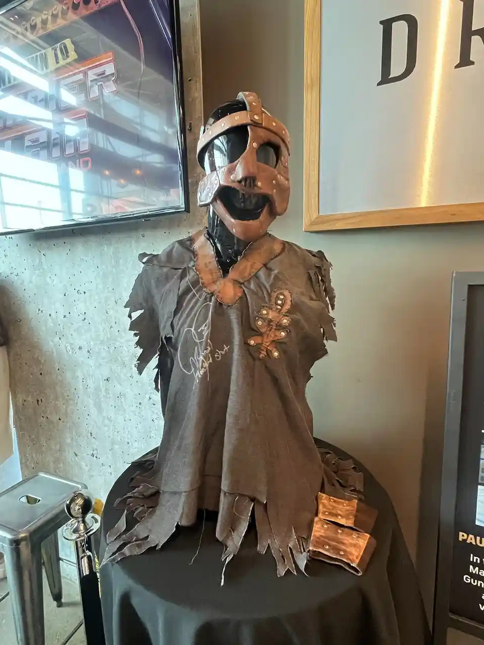 A mannequin wearing a brown leather mask with a wide-open mouth, a torn grey t-shirt with a signature and a brown leather…