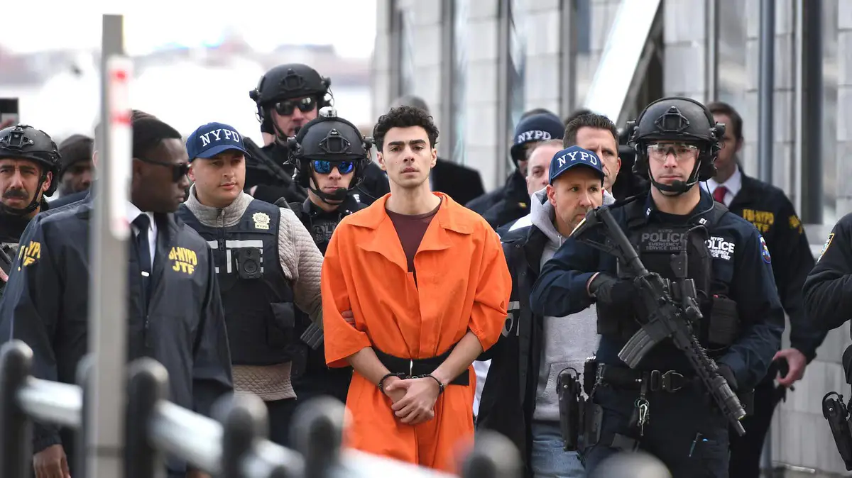 A man in an orange prison jumpsuit stands handcuffed, surrounded by heavily armed NYPD and FBI agents in tactical gear. The…