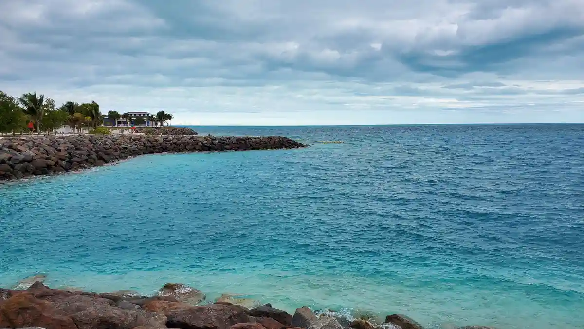 A scenic photo of a calm tropical beach with turquoise water, a rocky breakwater, palm trees, and a building in the distance…