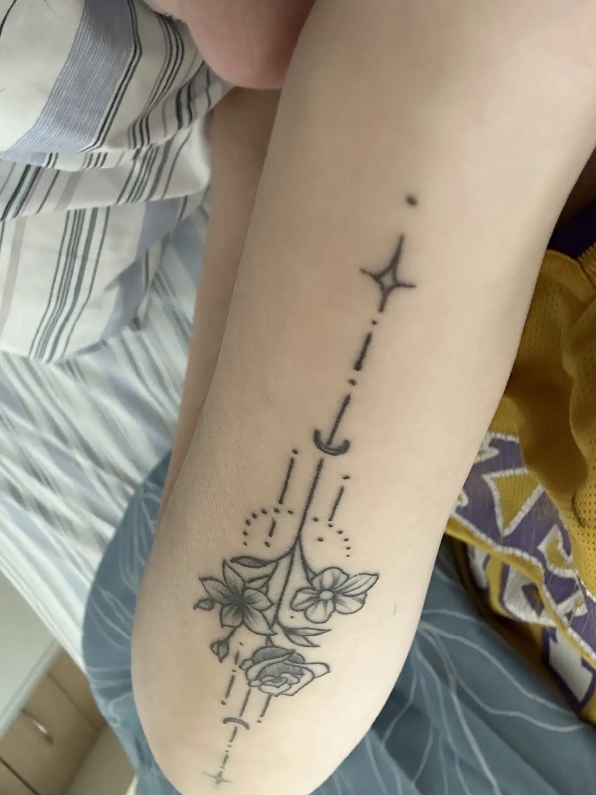 A close-up photo shows a person's forearm with a black ink tattoo featuring stylized flowers, crescent moons, and stars…