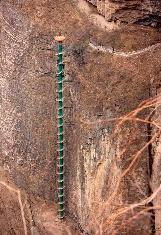 a vertical green glass walkway clinging to a sheer cliff face, with tiny figures of people walking on it, and a winding path…