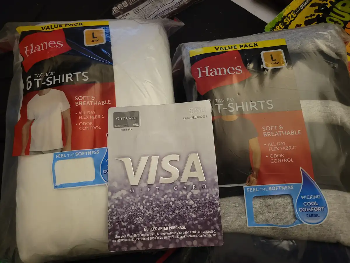Two packages of Hanes brand tagless t-shirts, one white and one gray, are displayed with a $50 Visa gift card placed between…
