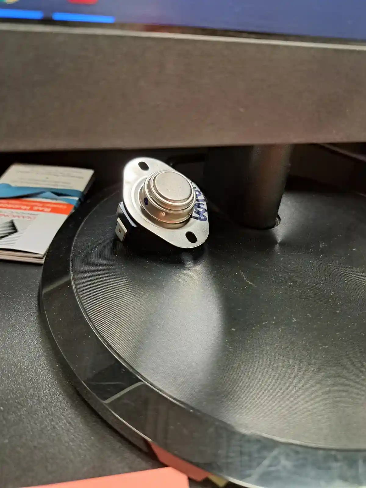 A close-up photo shows a silver-colored electronic component, a thermistor, resting on the black, textured base of a monitor…