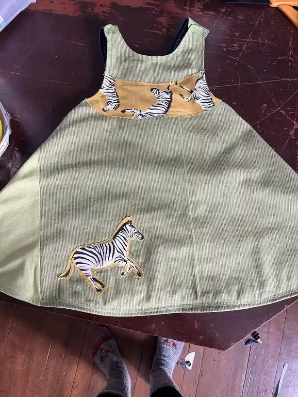 A handmade green dress with a zebra appliqué on the skirt and a zebra-patterned fabric band at the neckline, laid flat on a…