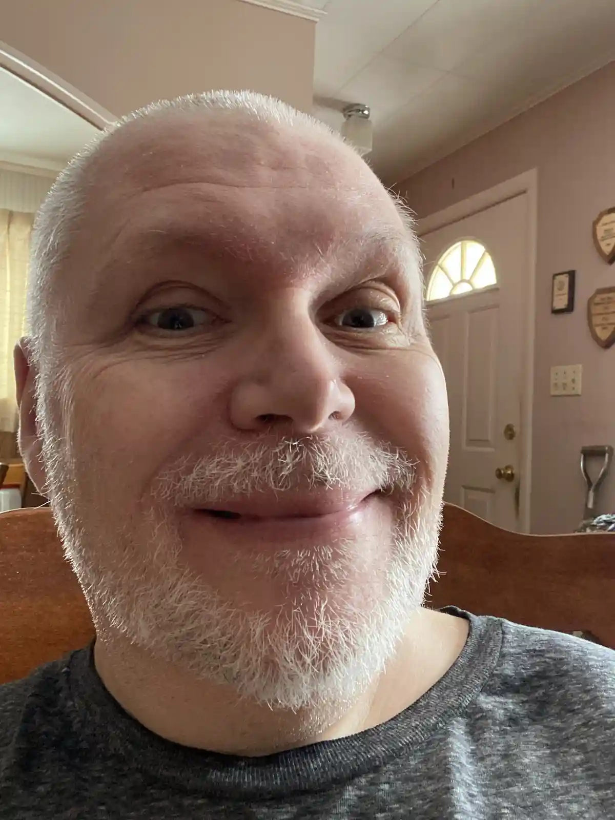 A close-up selfie of a middle-aged man with a shaved head and white beard, smiling with wide eyes while seated indoors. The…