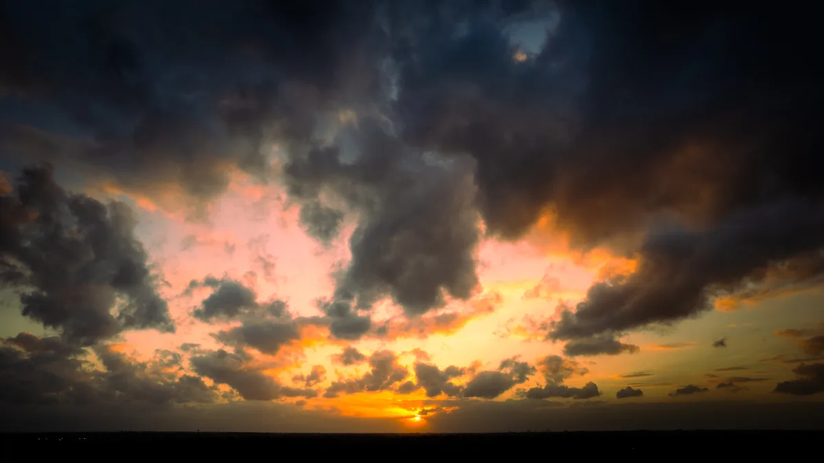 A dramatic sunset with dark, brooding clouds tinged with fiery orange and pink hues, the sun low on the horizon. Overlaid…