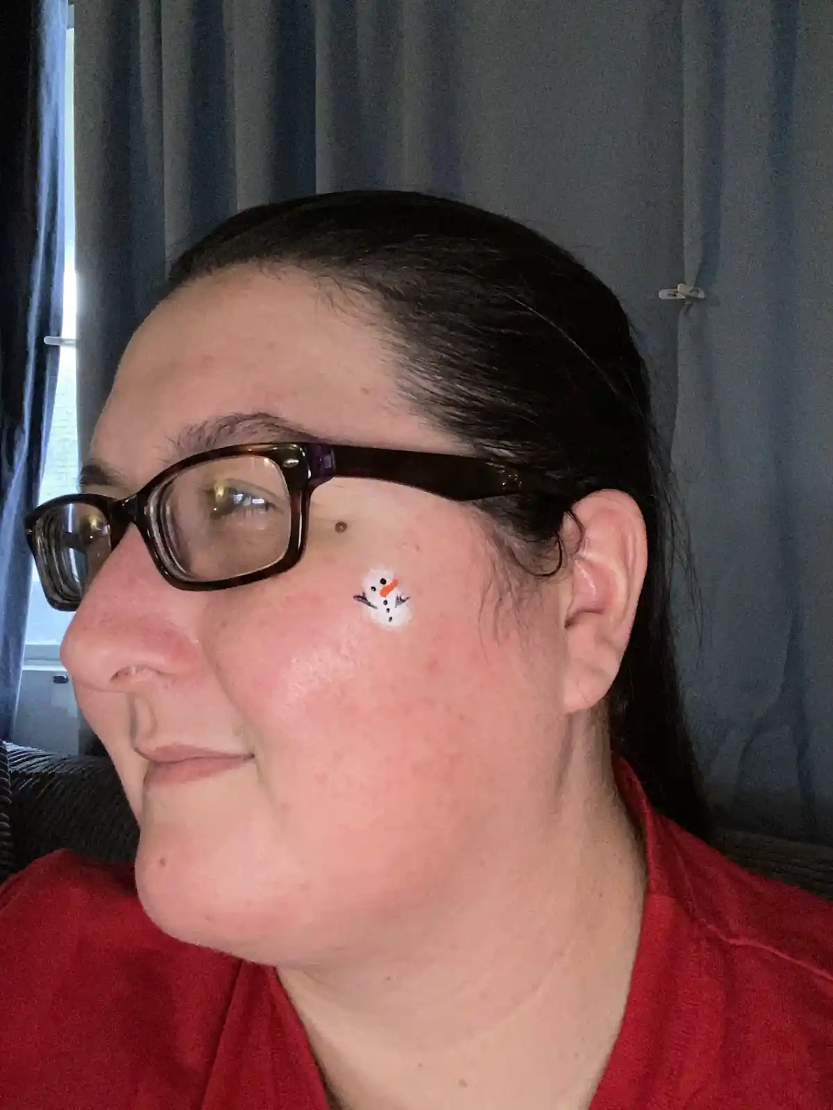 A person with dark hair and glasses, wearing a red shirt, has a small white snowman drawn on their cheek with black and…