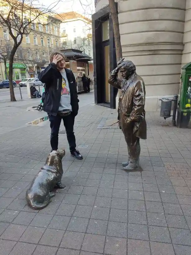A man stands on a city sidewalk next to a bronze statue of a man in a similar pose, both with one hand on their head. A…