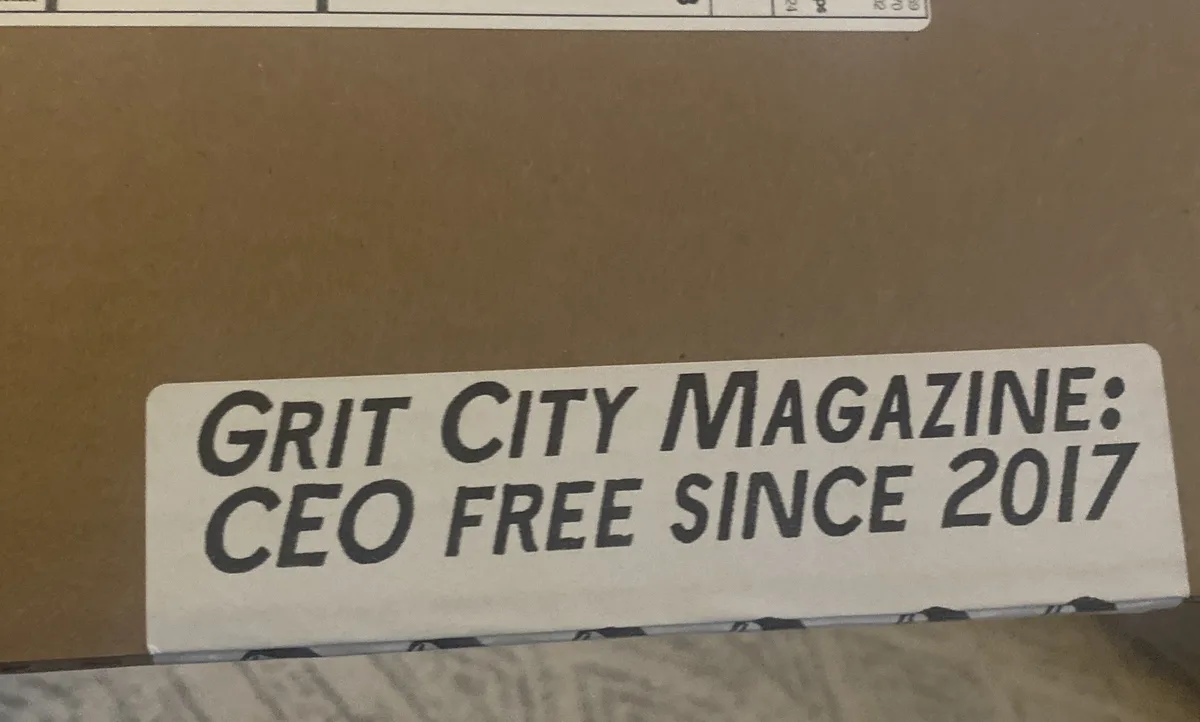 A close-up photo shows a white rectangular sticker on a brown cardboard box. The sticker has bold, black, sans-serif text…