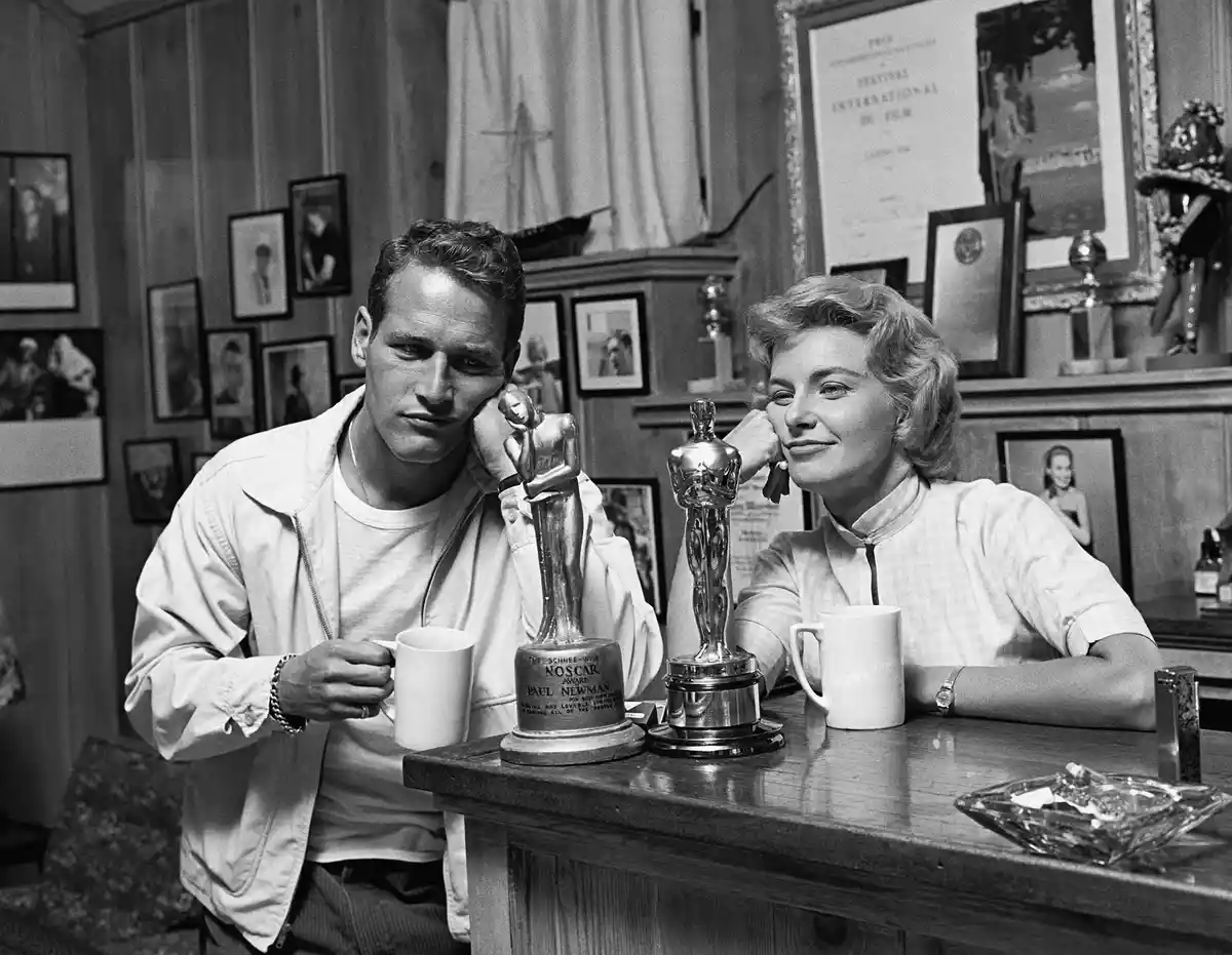 A black-and-white photo shows actors Paul Newman and Joanne Woodward sitting at a wooden table with two trophies between…