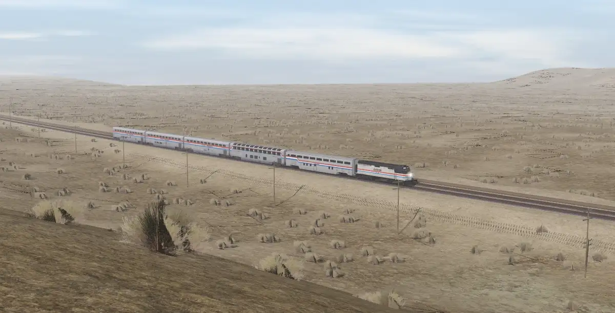 A long Amtrak passenger train travels through a vast, arid desert landscape with sparse vegetation and rolling hills under a…