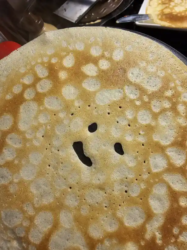 A close-up photo of a golden-brown pancake with numerous bubbles on its surface, which has been edited to show a sad face…