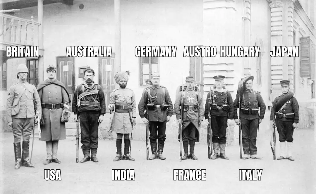 A historical black-and-white photo shows seven soldiers in early 20th-century military uniforms standing in a line. Each…