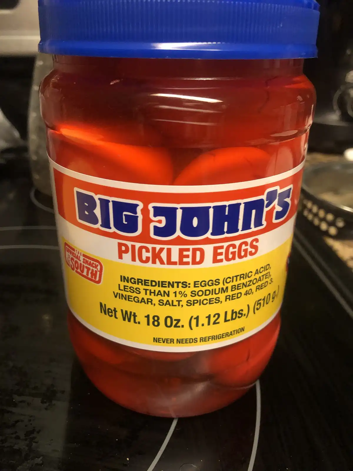 A large plastic jar with a blue lid contains bright red pickled eggs submerged in brine. The label reads 'BIG JOHN'S PICKLED…