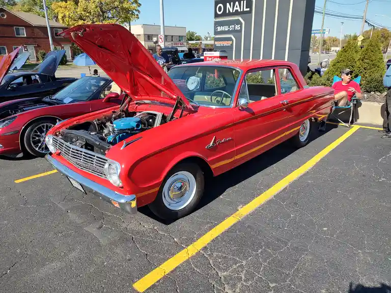 A red vintage Ford Falcon with its hood open is parked in a lot with other cars, while a man in a red shirt sits in a…