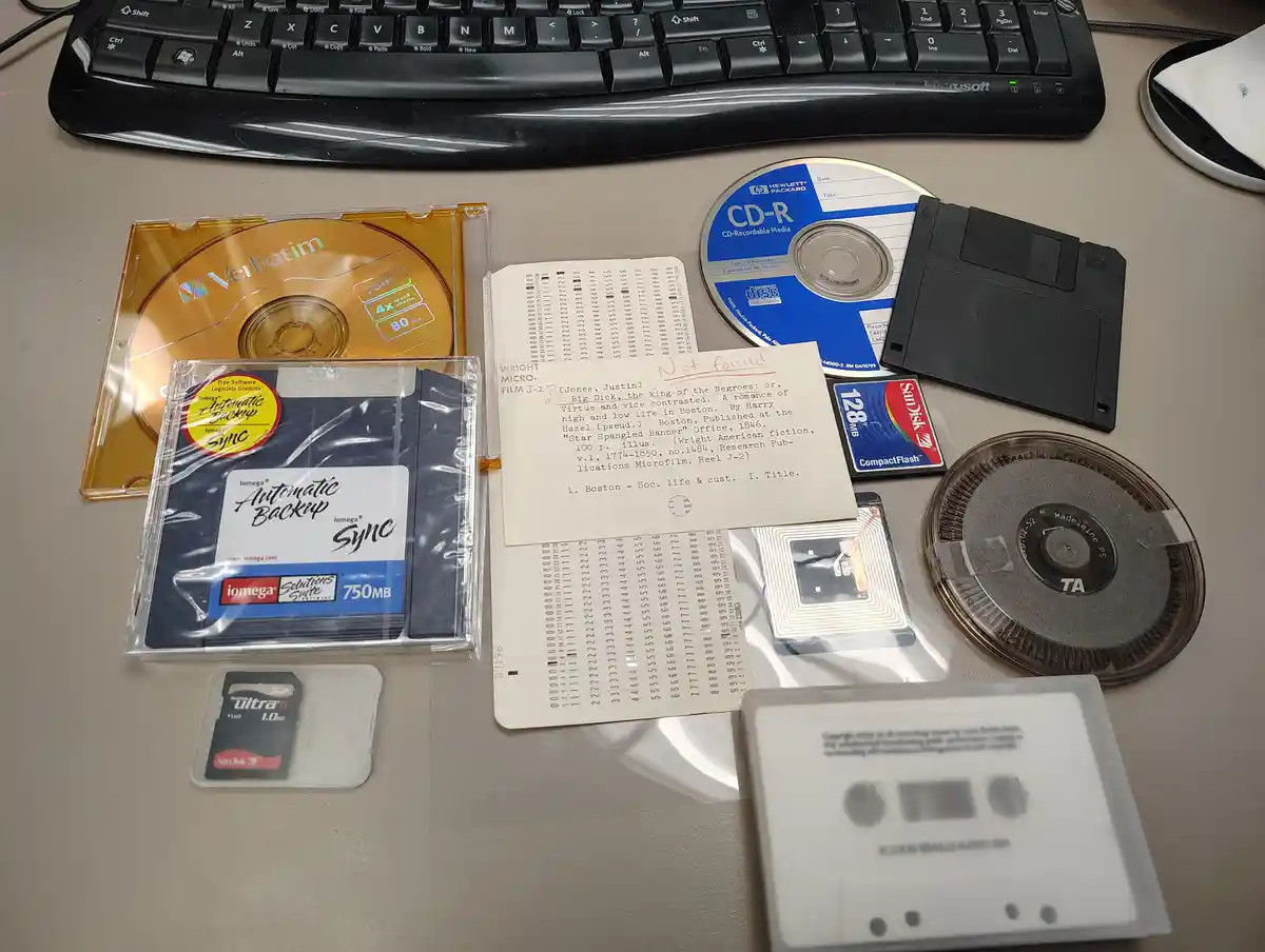 An overhead shot displays a collection of obsolete computer storage media on a desk, including a Verbatim CD-R, an Iomega…