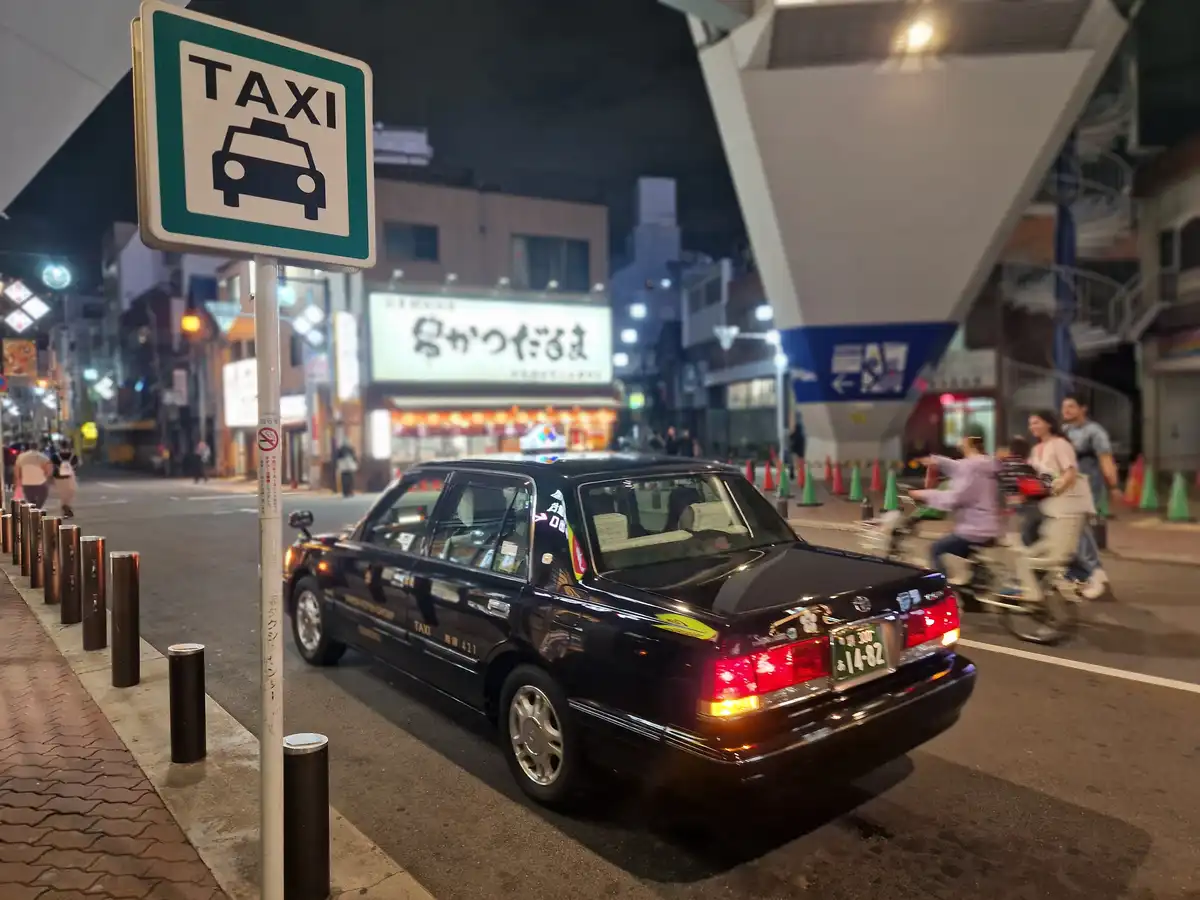 A black taxi is parked at night beside a green and white 'TAXI' sign on a city street, with blurred pedestrians and cyclists…