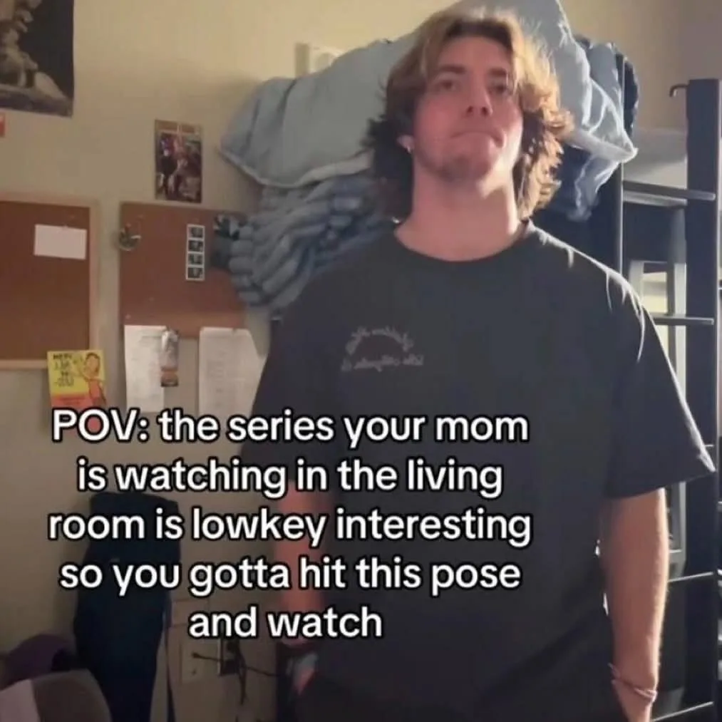 A young man with shoulder-length hair stands in a cluttered room, looking upward with a bored or resigned expression. Behind…