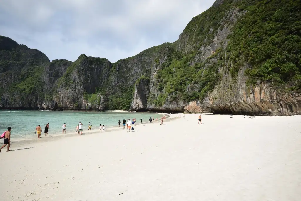 a wide-angle photo of a stunning tropical beach with white sand, turquoise water, and towering green-covered cliffs. Several…