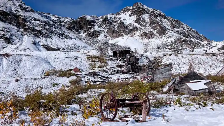 A snowy, desolate landscape featuring abandoned wooden buildings and a rusted wagon wheel in the foreground, set against a…