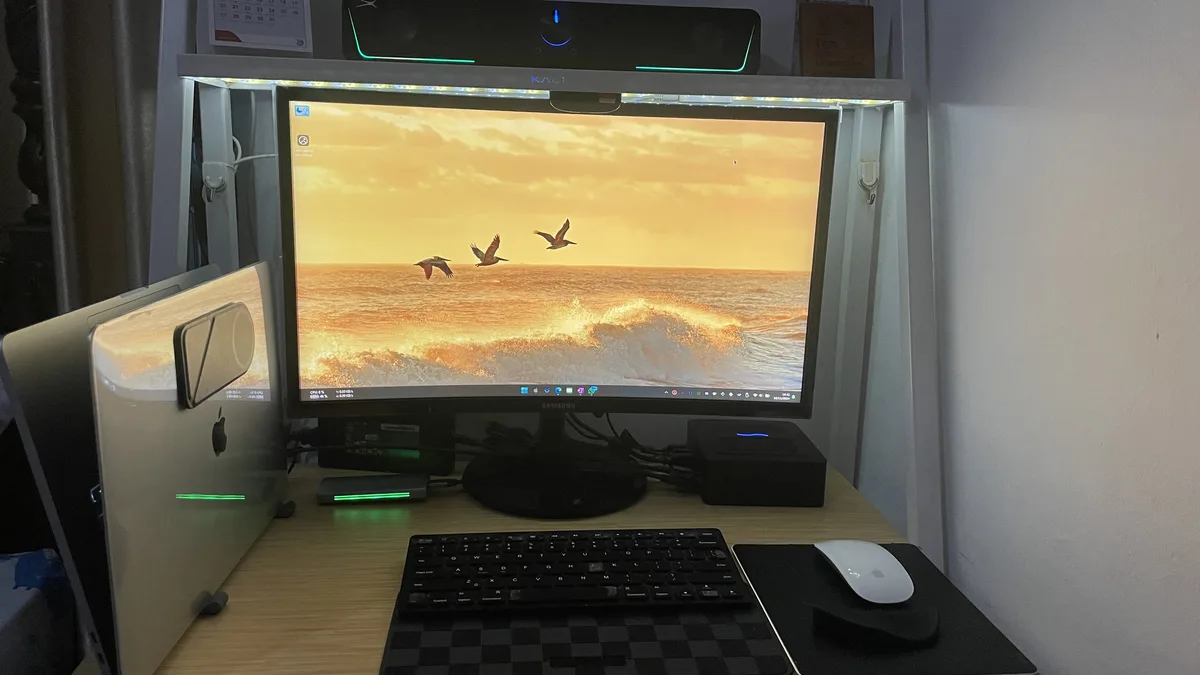 A computer desk setup with a Samsung monitor displaying a sunset wallpaper of birds flying over ocean waves, next to an…