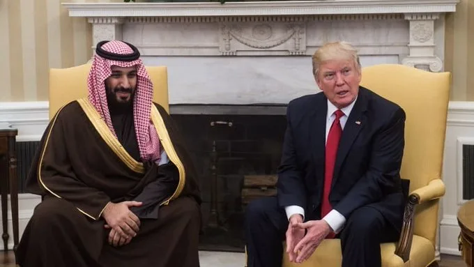 Two men sit in yellow chairs in front of a fireplace. The man on the left wears traditional Saudi attire including a…