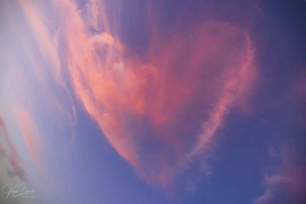 A photograph of a sky with a large, soft cloud formation shaped like a heart, glowing with pink and orange hues against a…