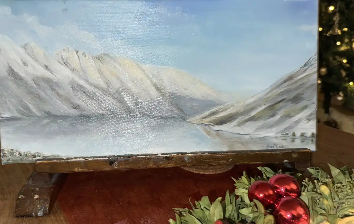 A painting of snow-covered mountains and a calm lake sits on a wooden easel, surrounded by Christmas decorations including…