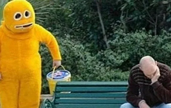 A person in a full-body yellow mascot costume holding a bucket stands next to a green park bench. On the bench, a bald man…