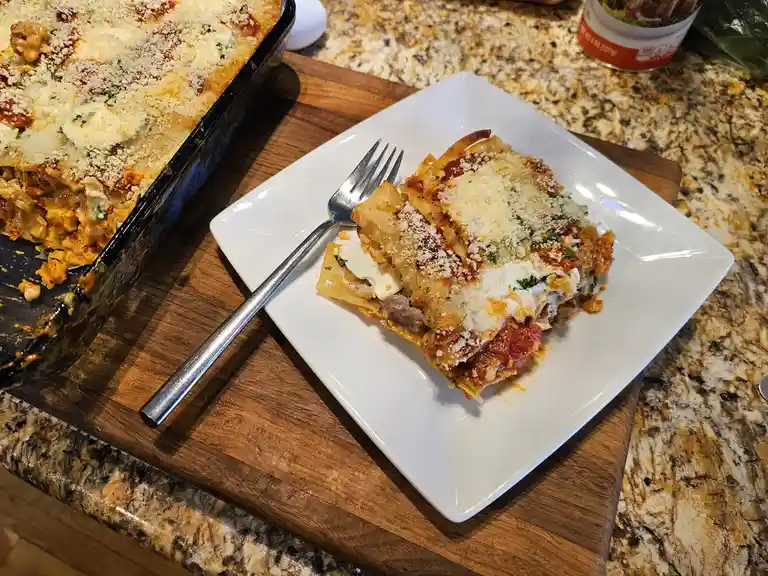 A slice of lasagna sits on a white plate with a fork, next to a baking dish from which it was served, on a wooden cutting…