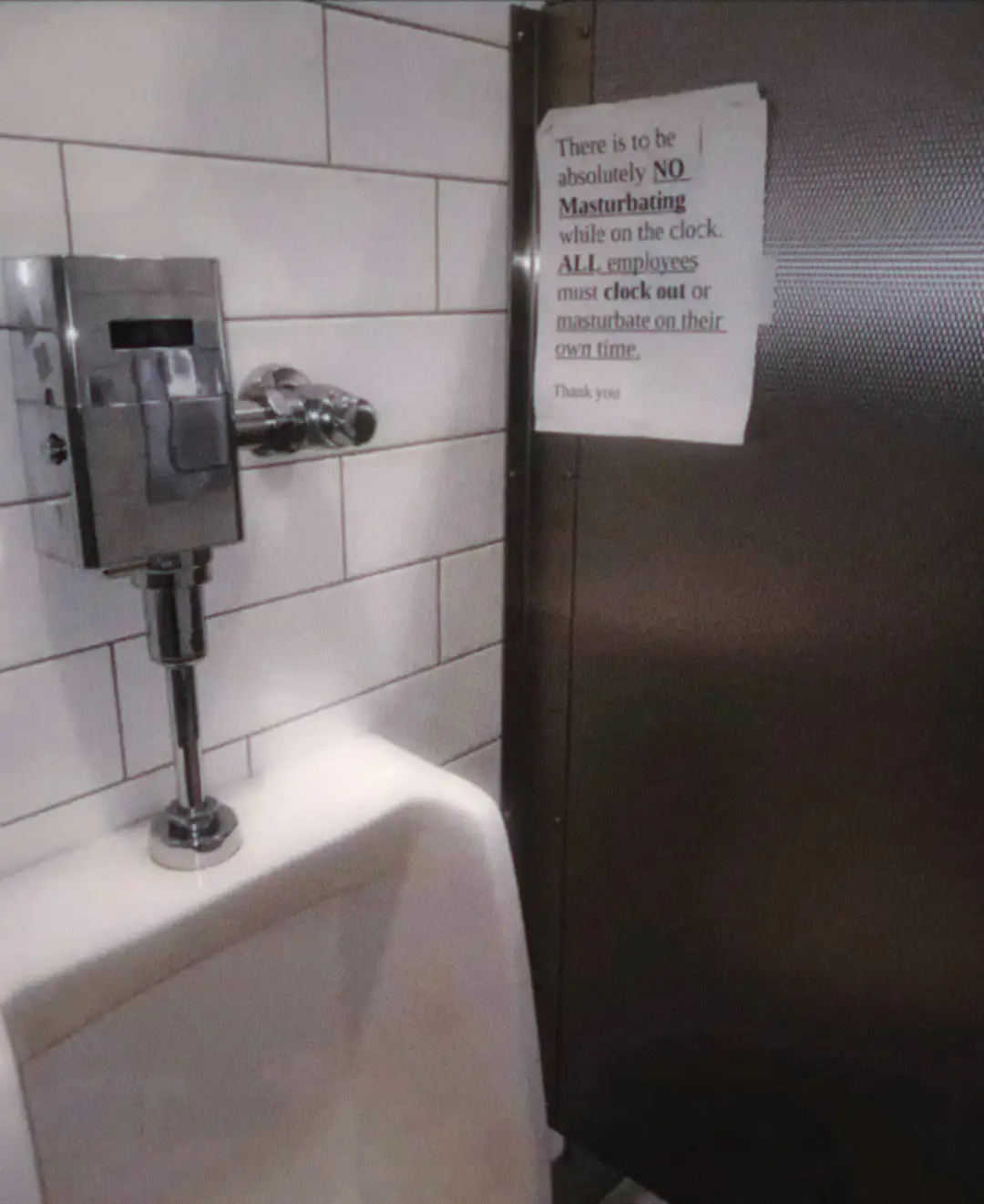 A photo of a public restroom urinal with a metal flush valve, next to a dark stall door. A white paper sign is taped to the…