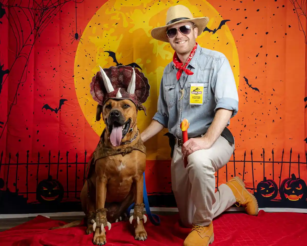 A man dressed as a Jurassic Park visitor with a straw hat, sunglasses, and a red bandana kneels beside a brown dog wearing a…
