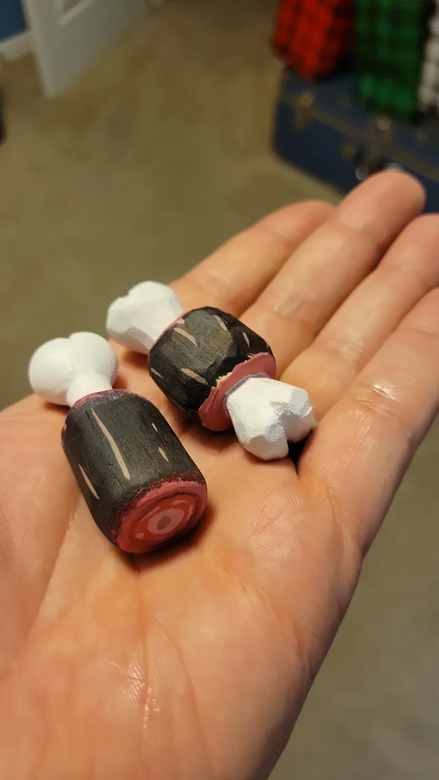A person's open palm holds two small, hand-painted wooden figurines resembling drumsticks, with black bodies, red ends, and…