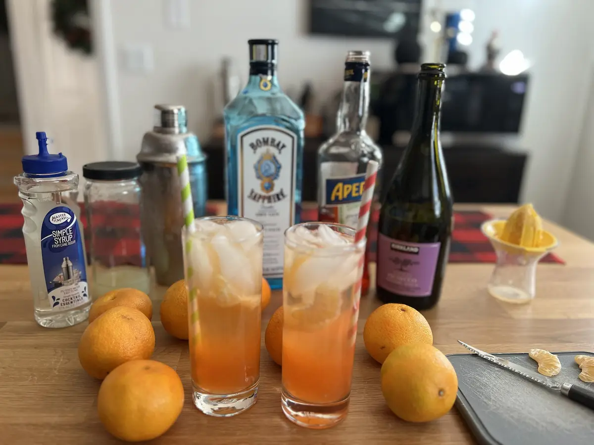 A wooden countertop displays two tall glasses filled with orange-colored cocktails and ice, surrounded by whole oranges, a…
