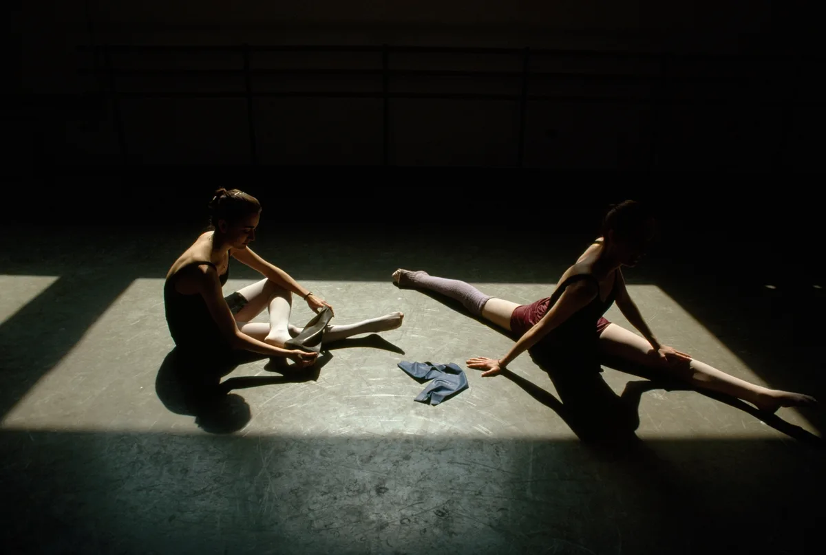 Two ballet dancers are in a dimly lit studio, illuminated by a single spotlight. One sits cross-legged, holding a ballet…