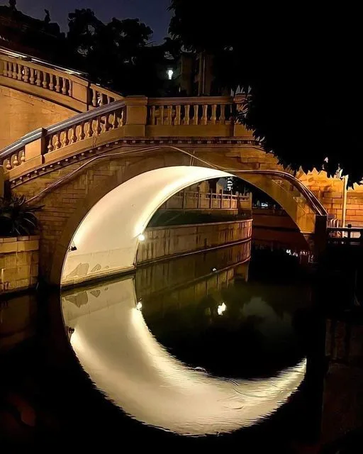 A nighttime photo of a stone arch bridge over a canal, where the illuminated underside of the bridge creates a perfect…
