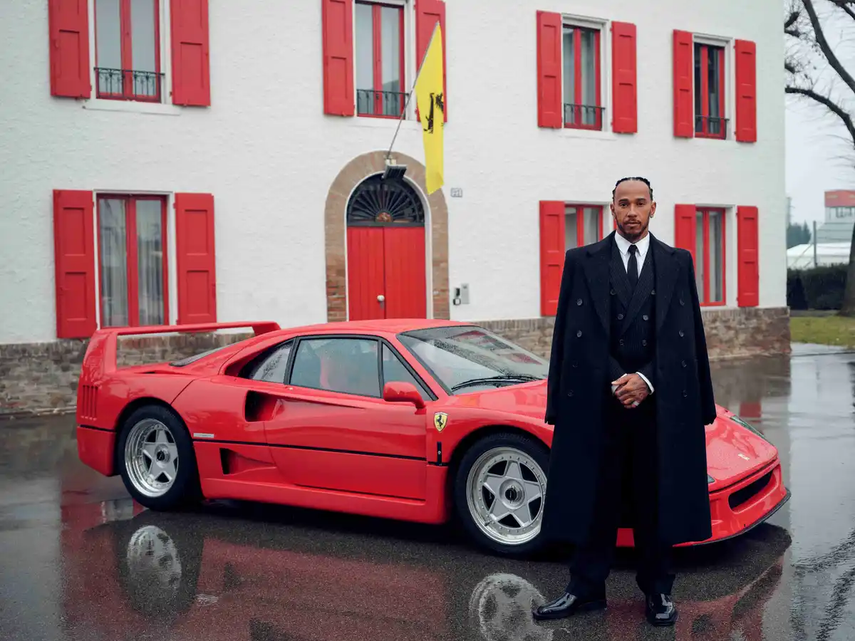 A man in a black double-breasted suit and long coat stands confidently next to a red Ferrari F40 on a wet pavement, with a…