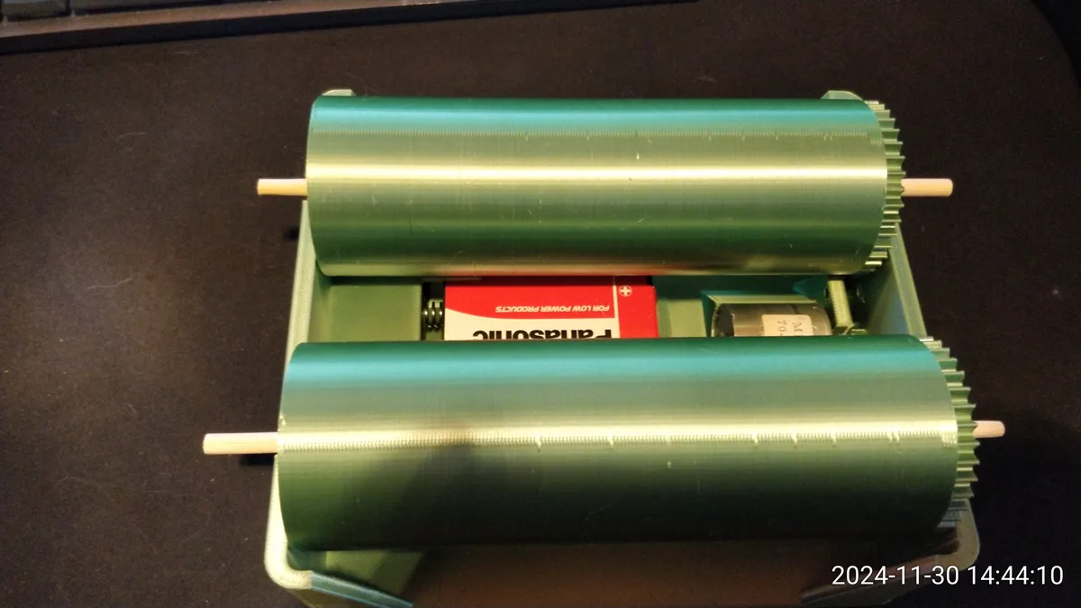 A close-up photo shows a green, open mechanical device with two large cylindrical rollers, revealing a red Panasonic battery…