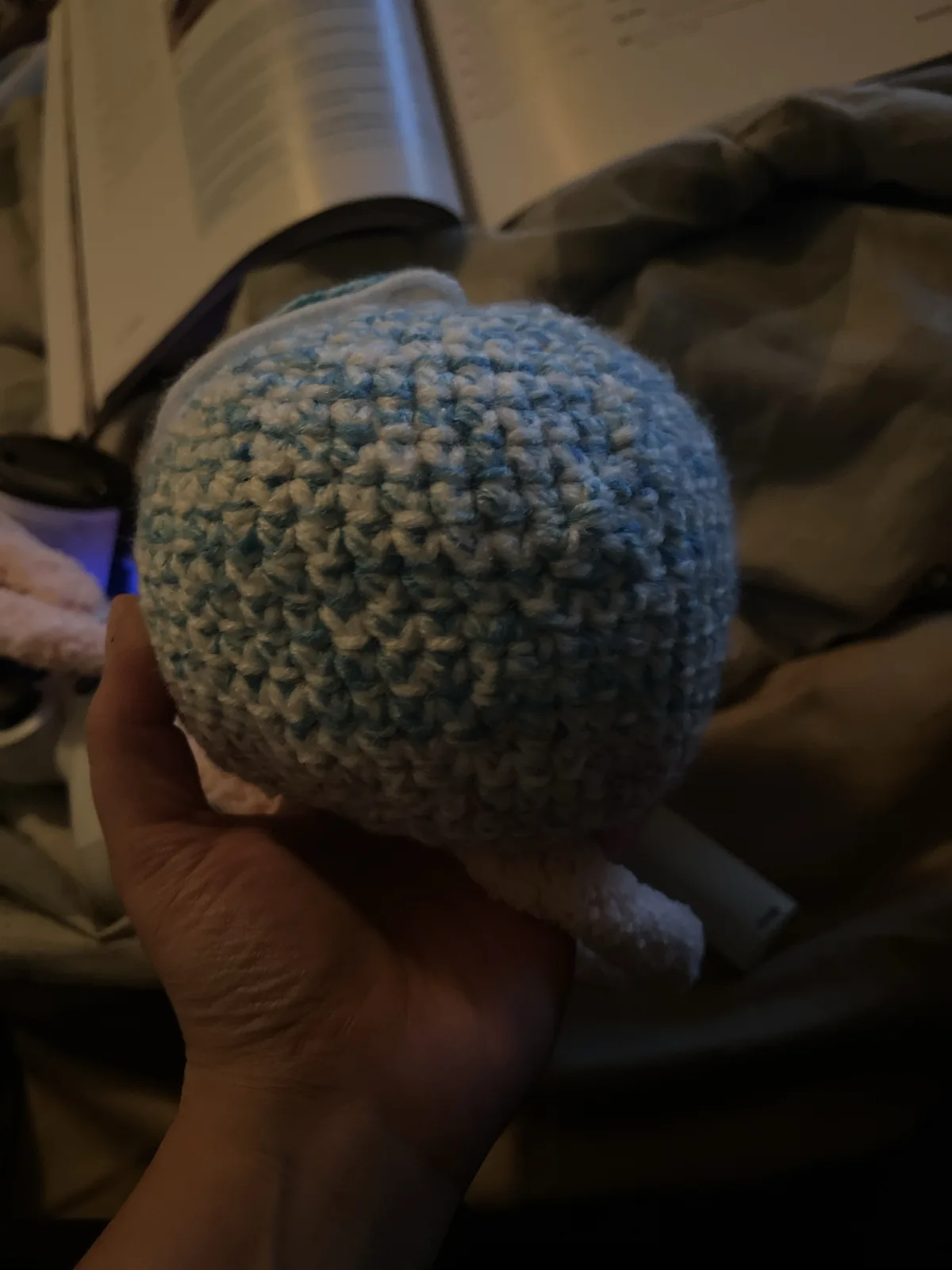 A hand holds a small, round, crocheted amigurumi toy made of light blue and white yarn, showing its backside with no facial…