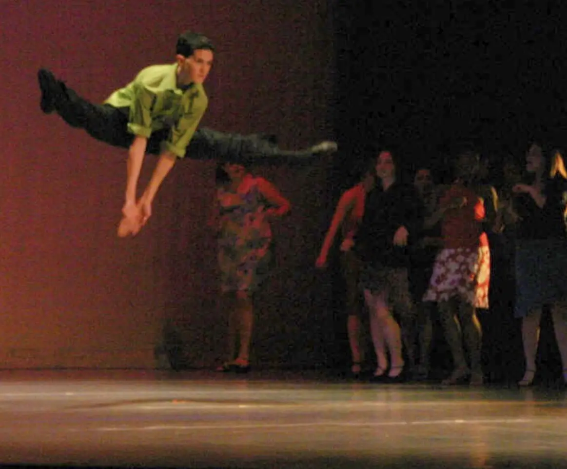 A young man in a green shirt and black pants is captured mid-air performing a split leap on a stage, with several blurred…