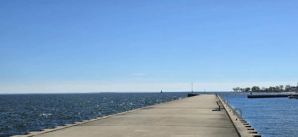 A wide-angle photo shows a long, empty concrete pier stretching into a vast blue body of water under a clear sky. The pier…