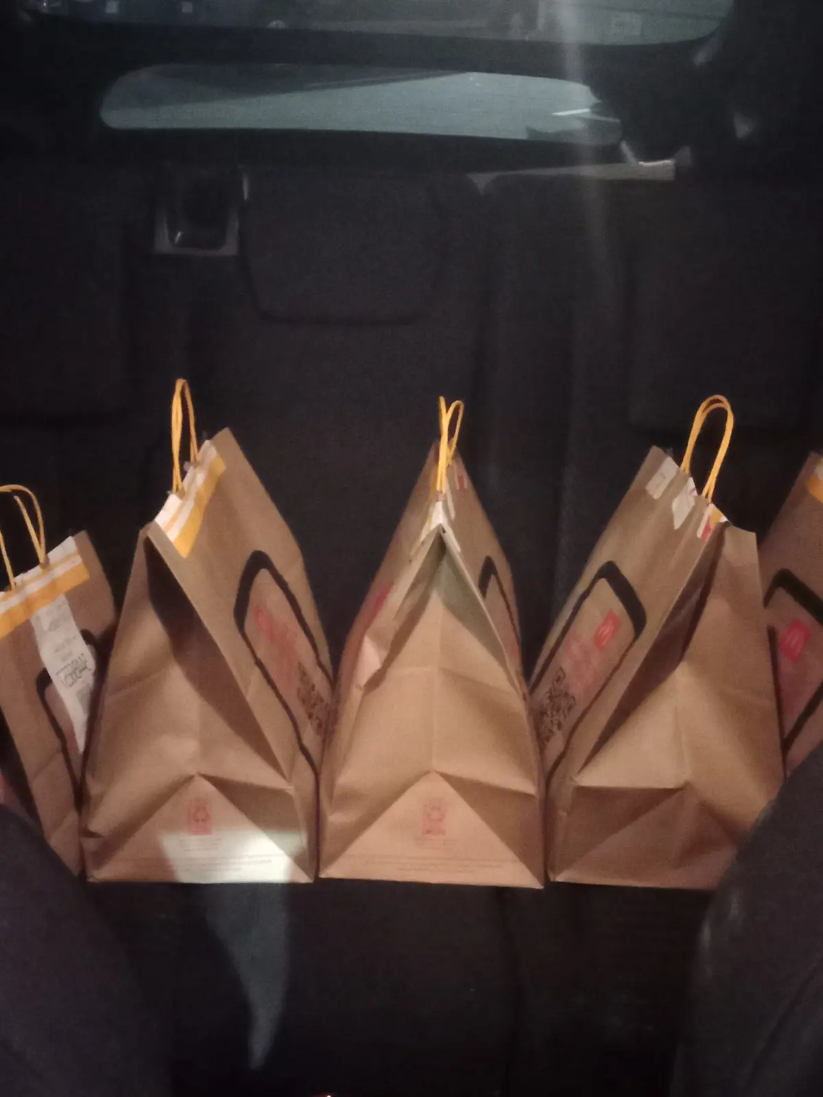 The image shows the back seat of a car filled with five brown paper bags from a restaurant, each hanging by its yellow…