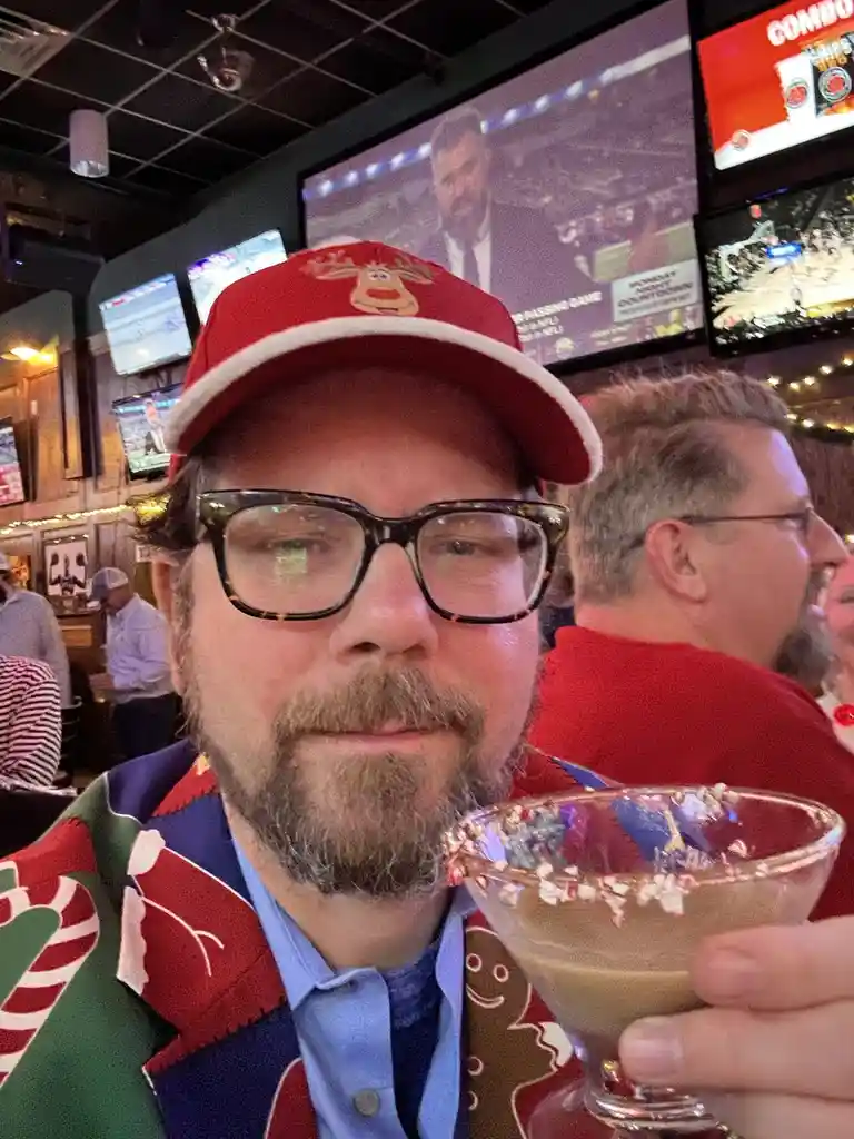 A man with a beard and glasses wears a red reindeer-themed cap and a colorful Christmas sweater with candy cane and…