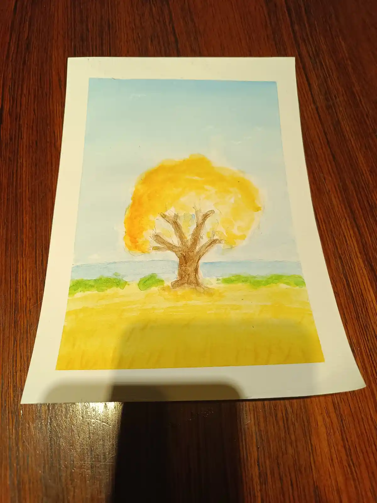 A watercolor painting on paper depicts a solitary tree with a bright yellow canopy and brown trunk, standing in a yellow…
