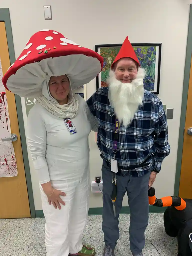 Two people pose indoors, one wearing a large red-and-white mushroom costume headpiece and a white uniform with an RN badge…