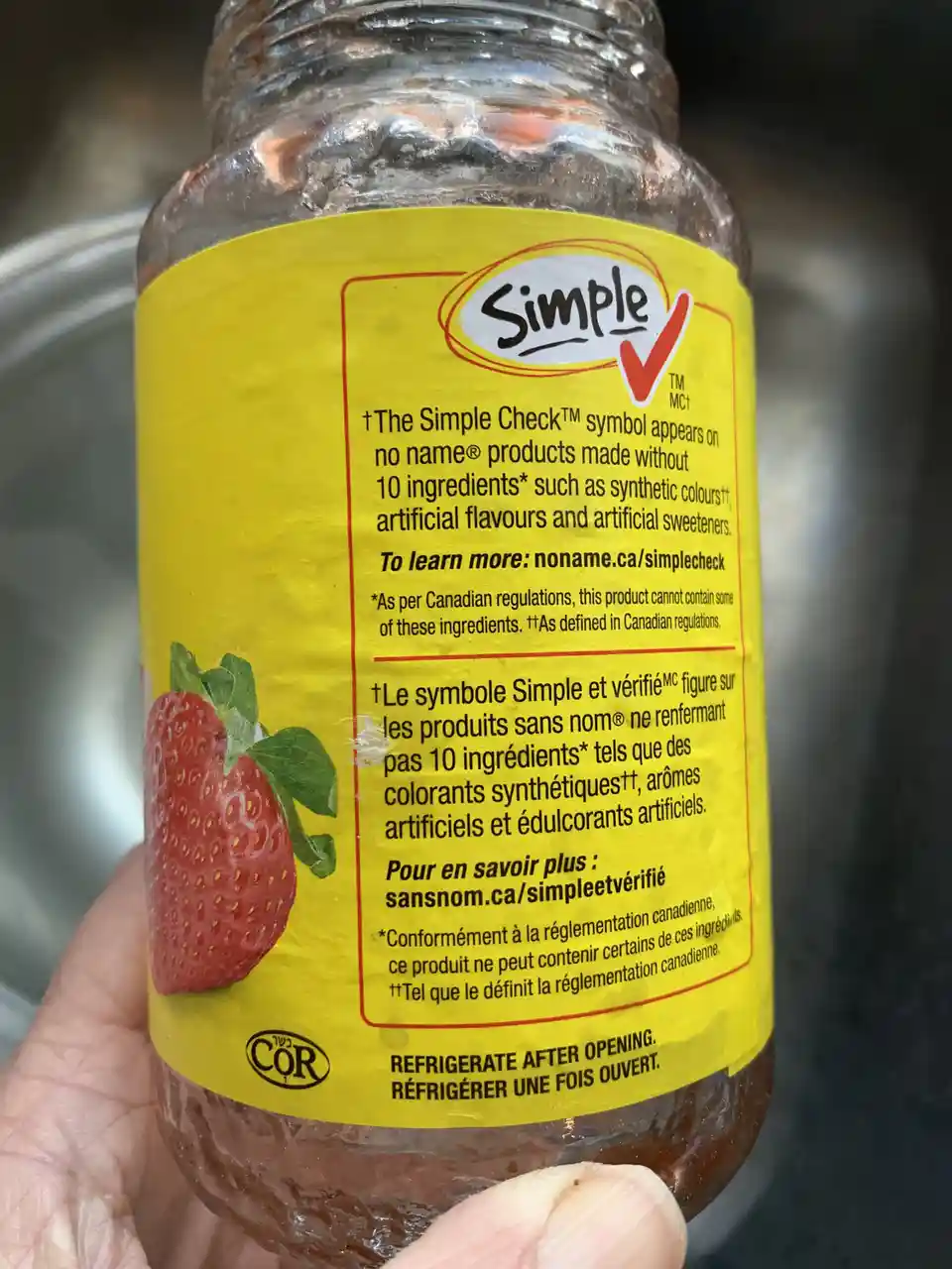A close-up photo shows a hand holding a glass jar of strawberry jam with a bright yellow label. The label features the…